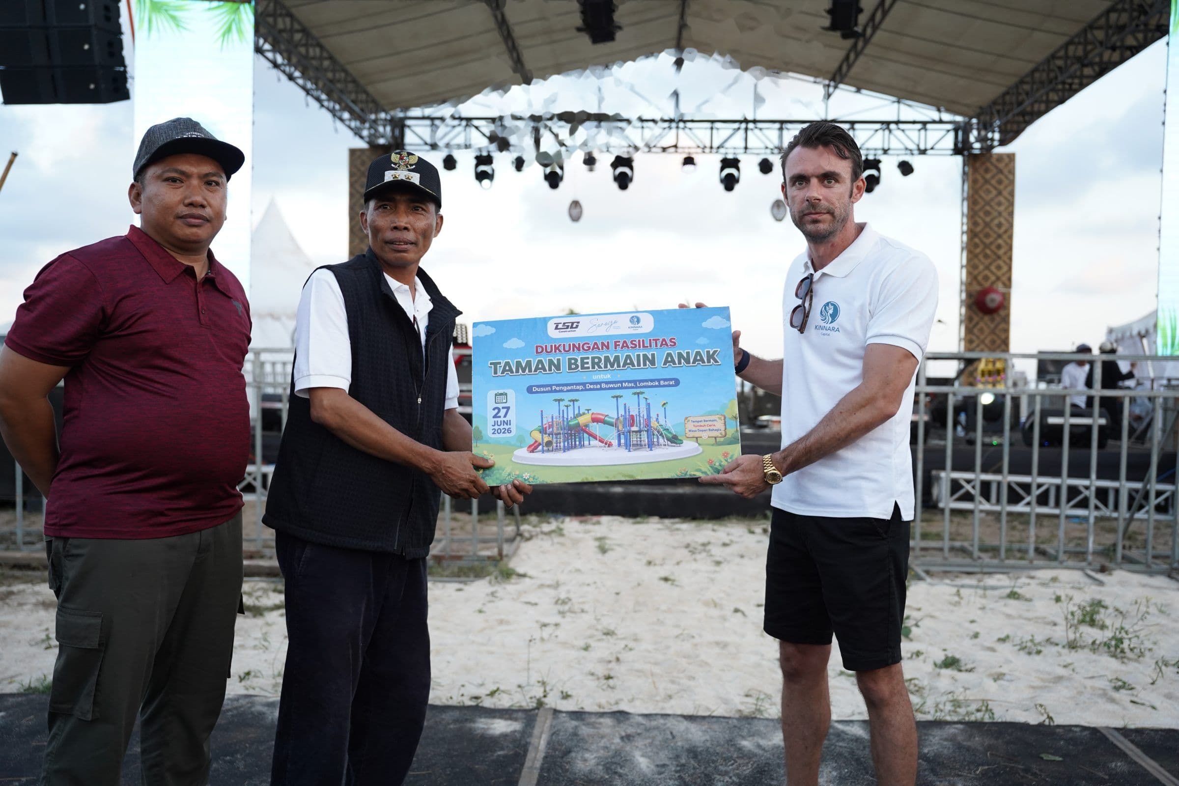 Adrian Campbell and two local officials holding a sign announcing support for a children's playground in Dusun Pengantap, Buwun Mas, in front of the beach stage.