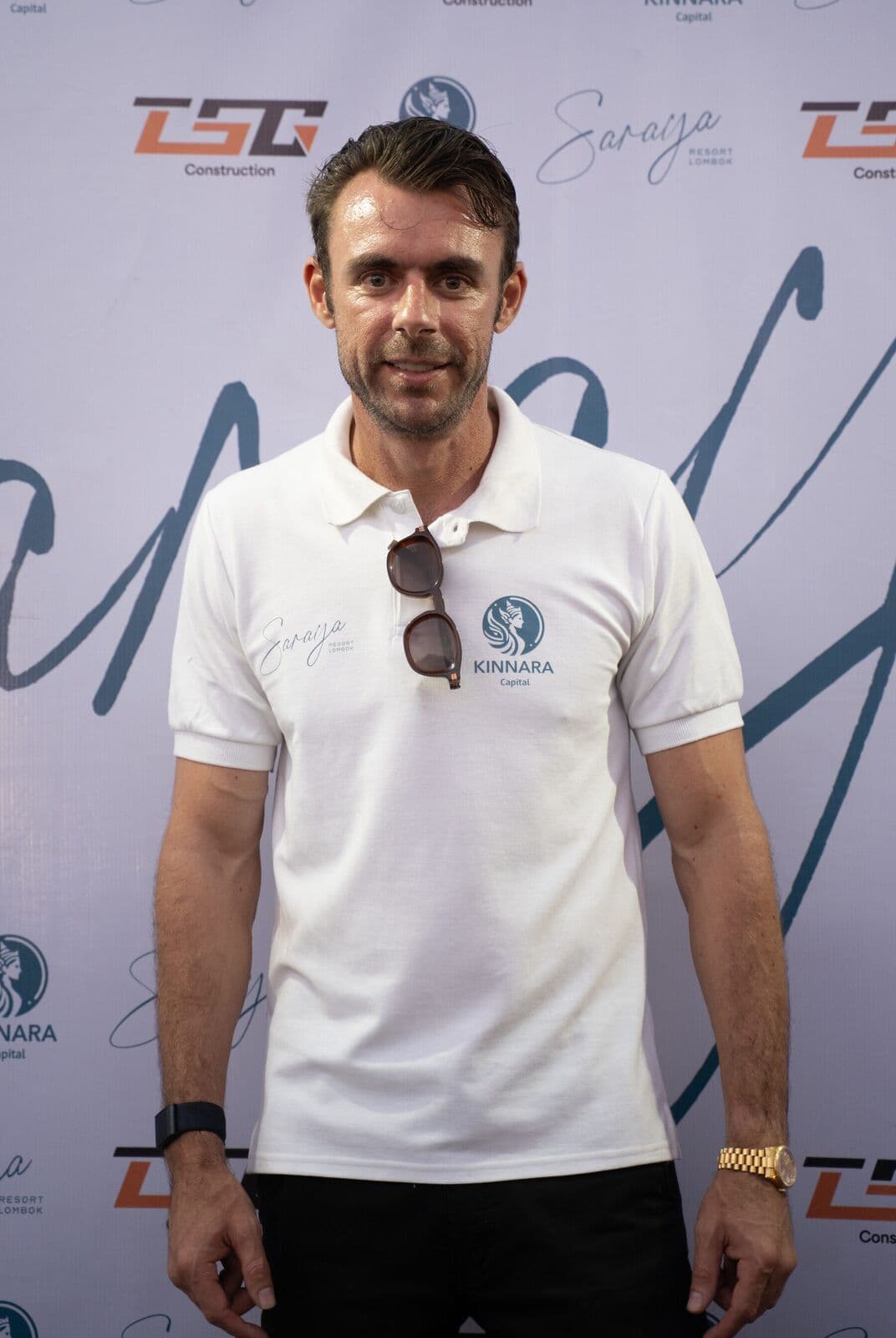 Adrian Campbell in a white Kinnara Capital polo shirt standing in front of the Saraya Beach Resort media wall at the Buwun Mas community day in West Lombok.