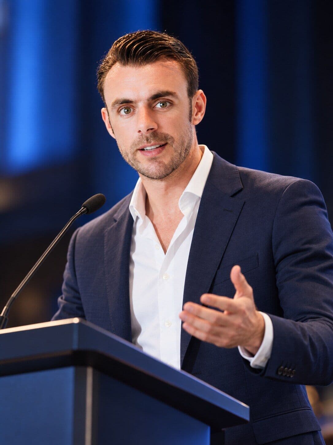 Adrian Campbell speaking at a lectern in a navy suit and open white shirt, gesturing with one hand, against a blue-lit background.