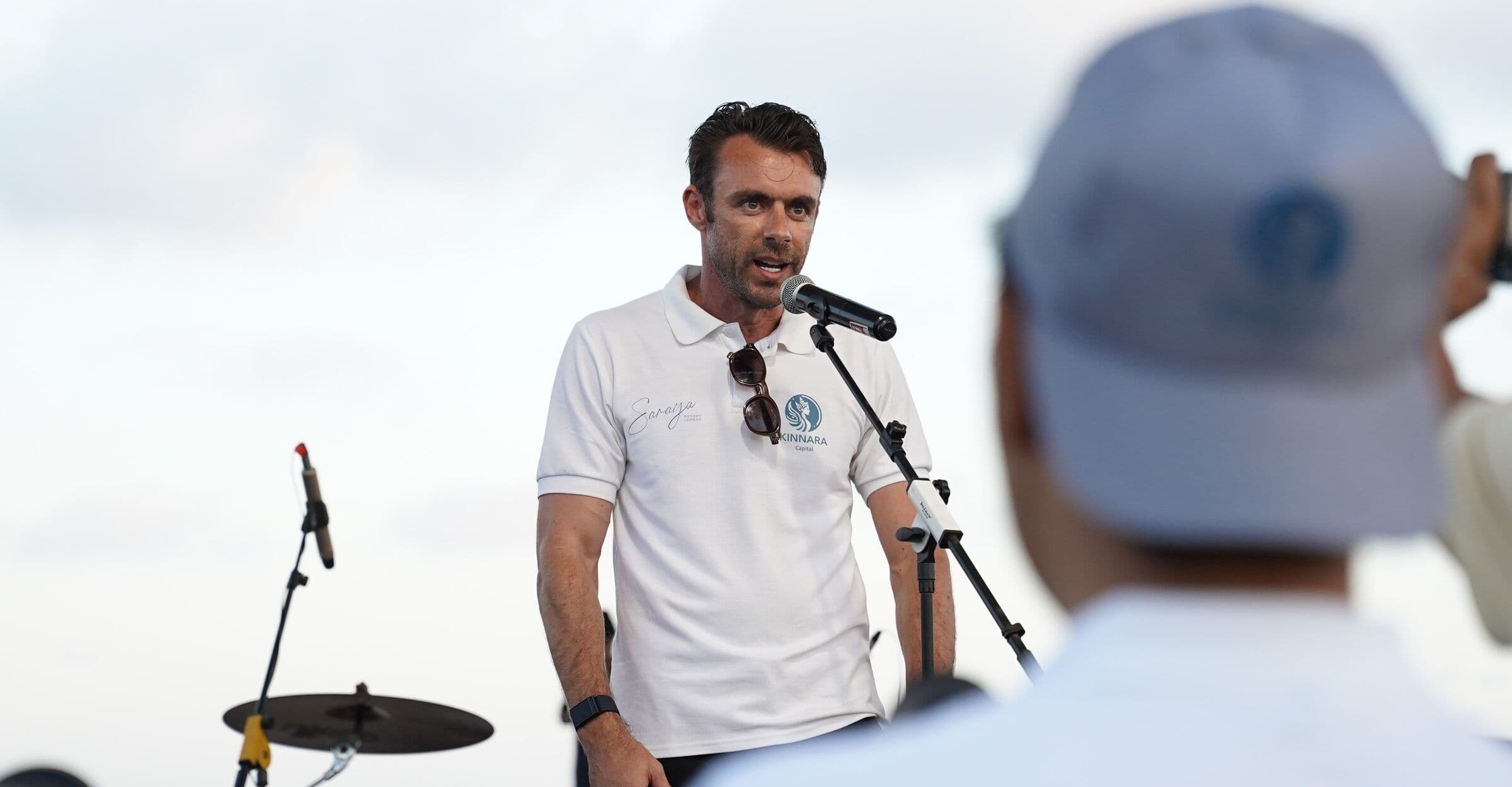 Adrian Campbell speaking at a microphone on the beach stage at the Saraya community day in Pengantap, Buwun Mas, Lombok, with villagers listening in the foreground.