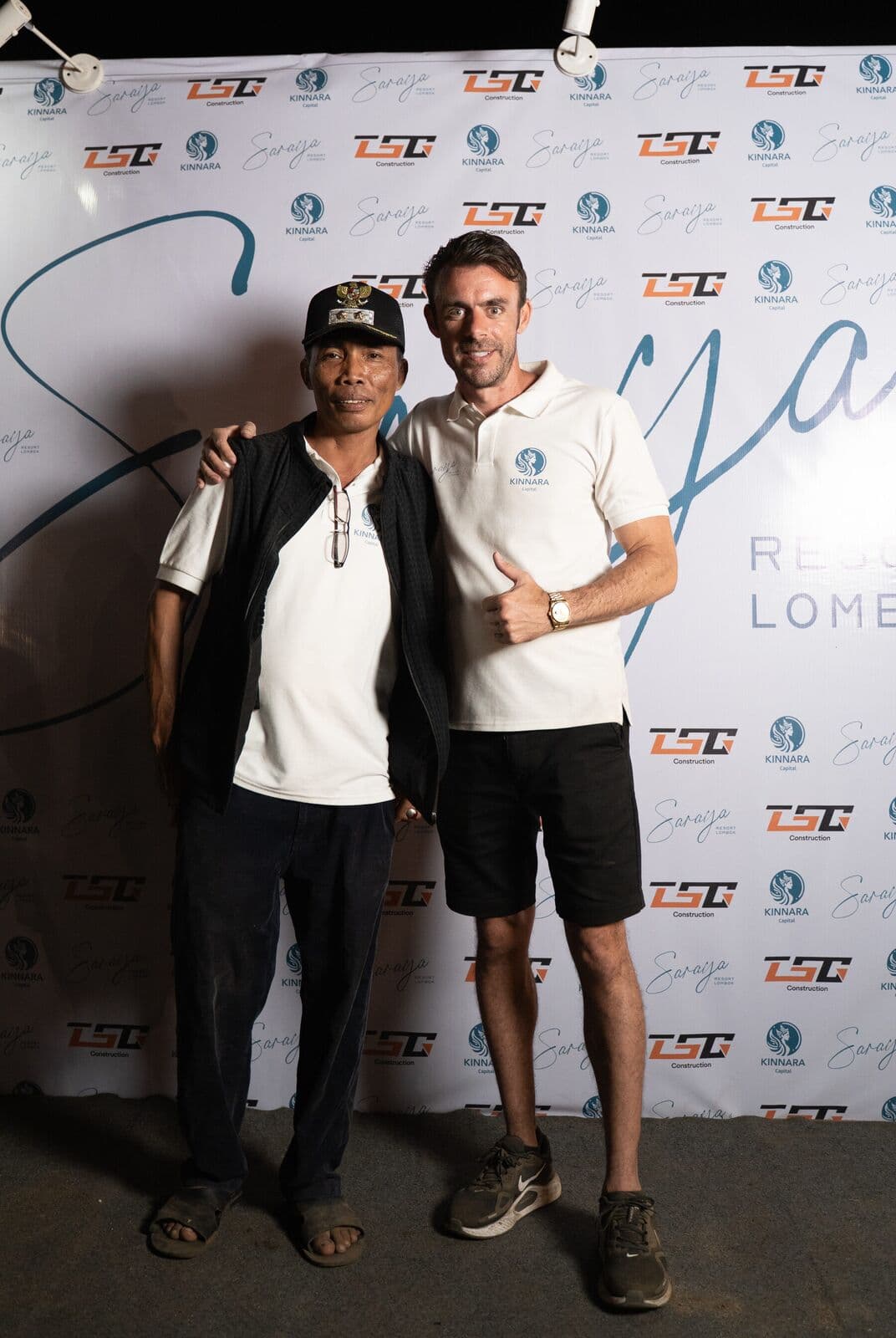 Adrian Campbell with his arm around a local Buwun Mas resident, both giving a thumbs up, in front of the Saraya Beach Resort and Kinnara Capital media wall.