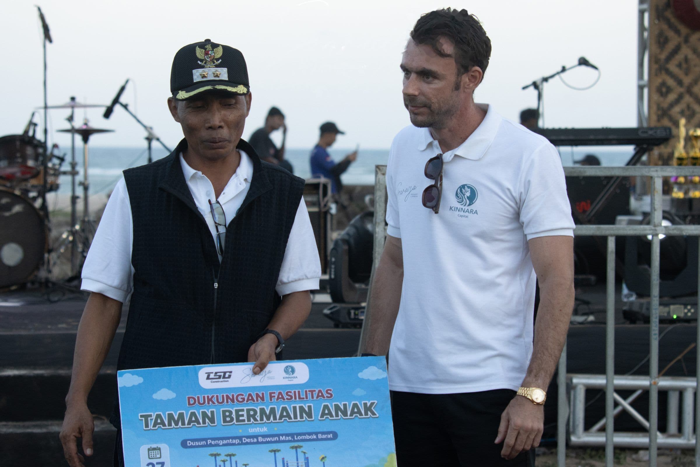 Adrian Campbell standing beside a local official in a peaked cap on the beach stage, the official holding the playground donation sign, with the sea behind.